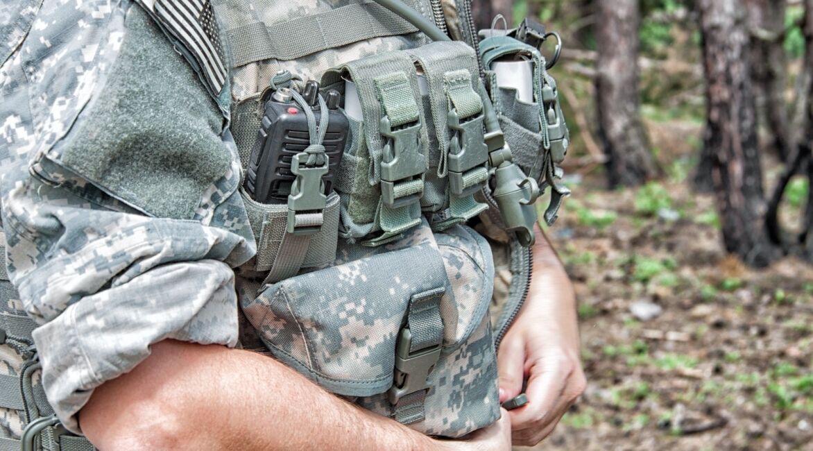 A close-up of a soldier's tactical vest featuring various pouches and equipment in a natural outdoor setting.