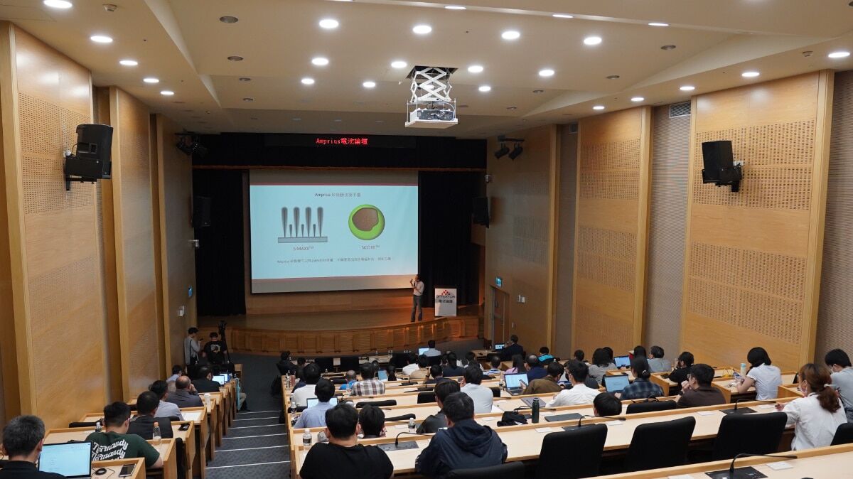 An auditorium filled with attendees, focused on a speaker presenting information on a large screen, displaying diagrams and graphs.