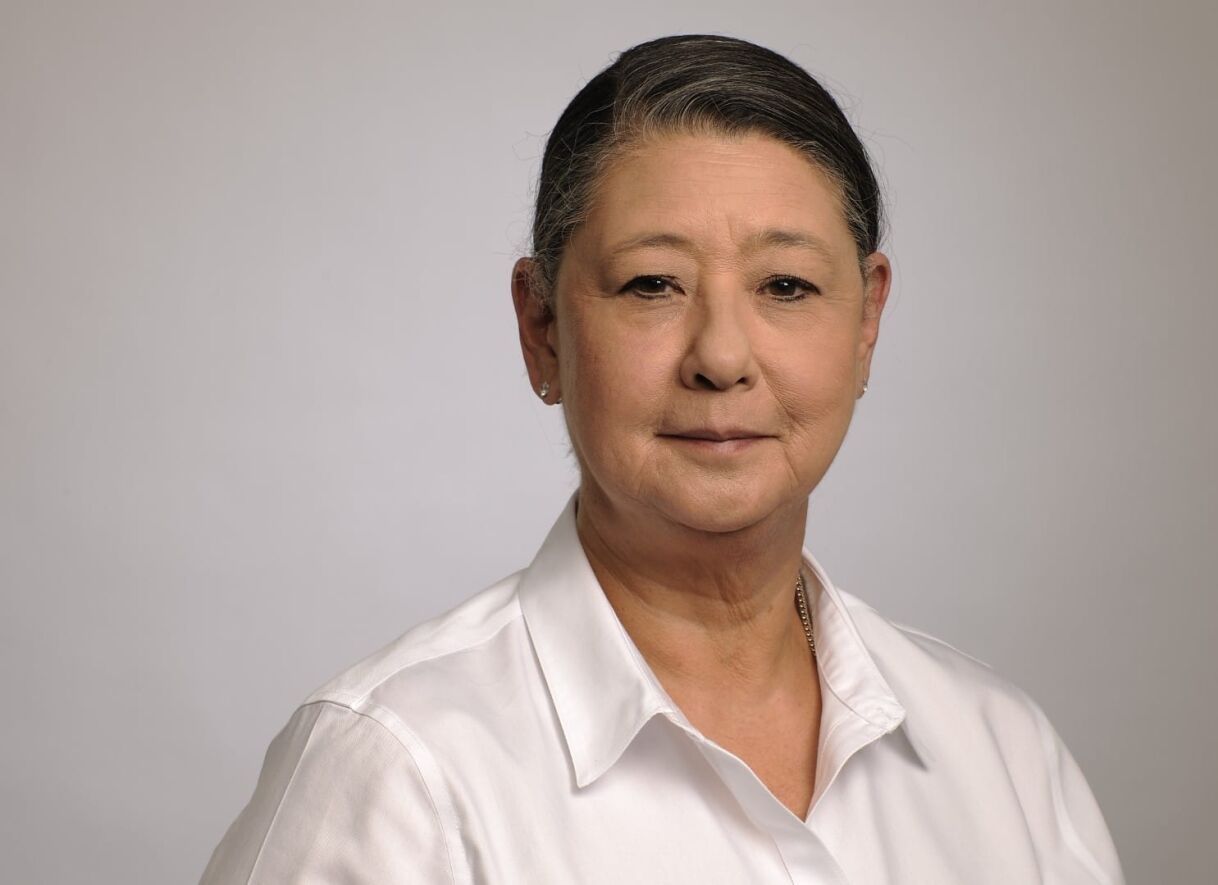 A woman with short gray hair, wearing a white shirt, looking directly at the camera with a neutral expression against a light background.