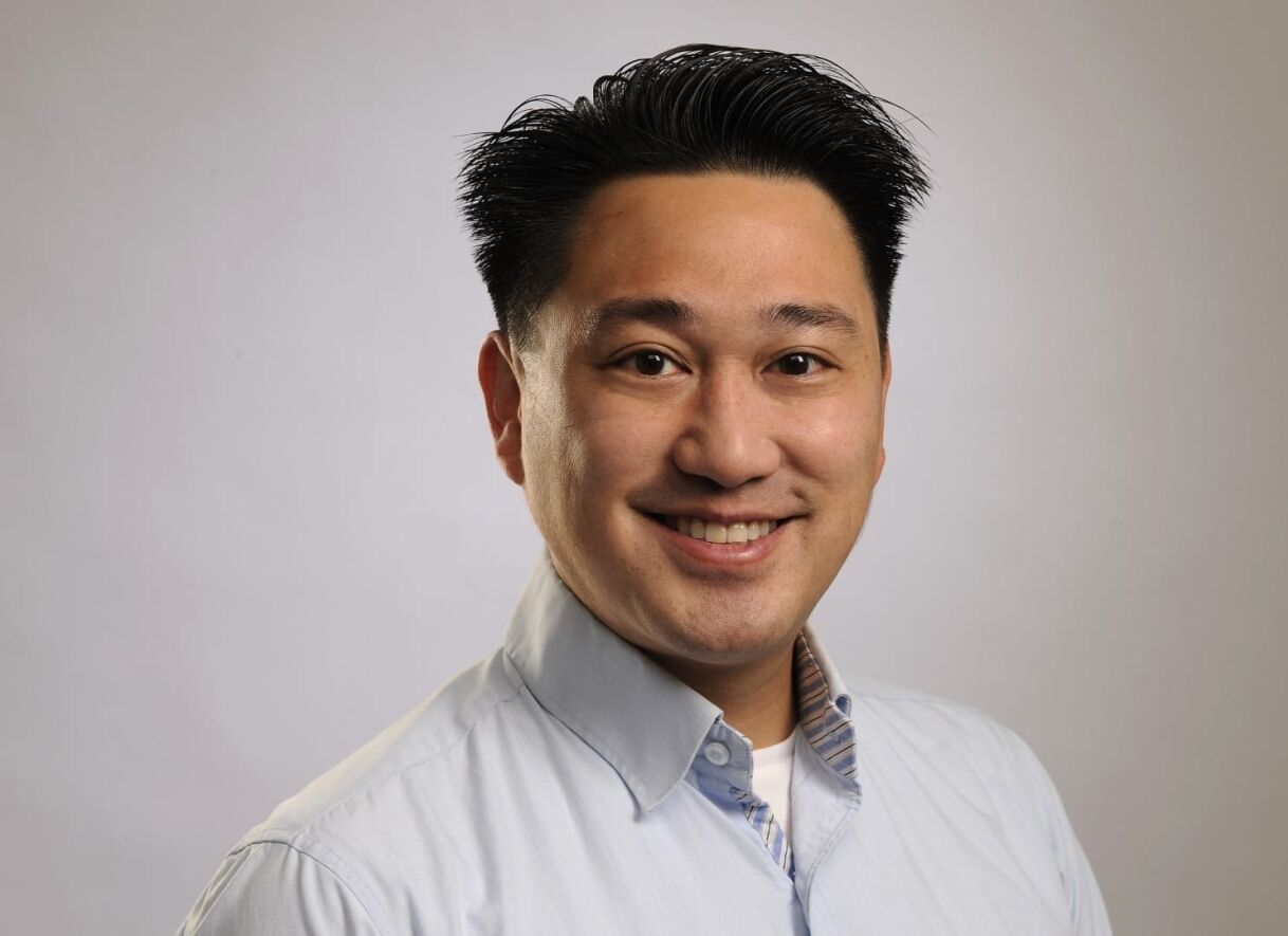 A smiling man with medium-length dark hair, wearing a light-blue collared shirt against a soft, neutral background.