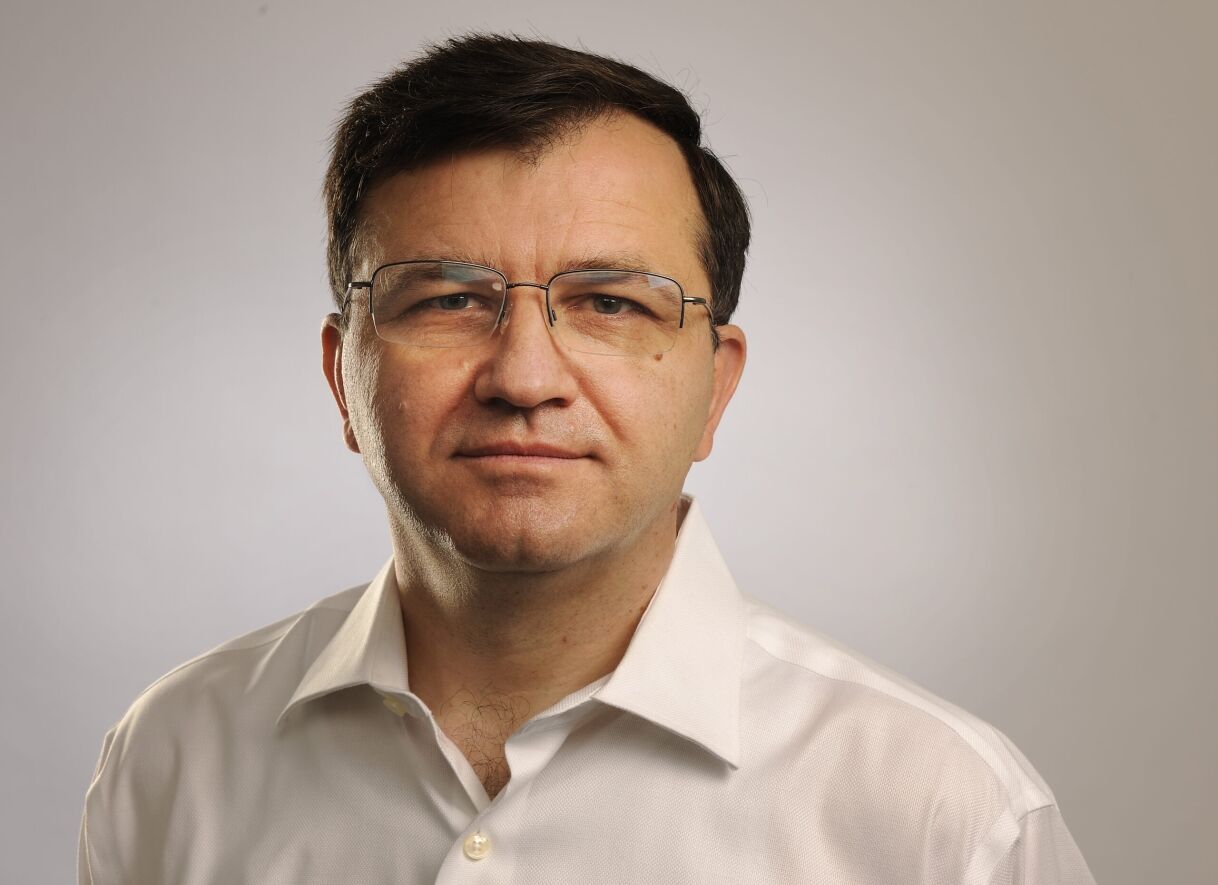 A man in a white shirt with glasses stands against a neutral background, presenting a serious expression.