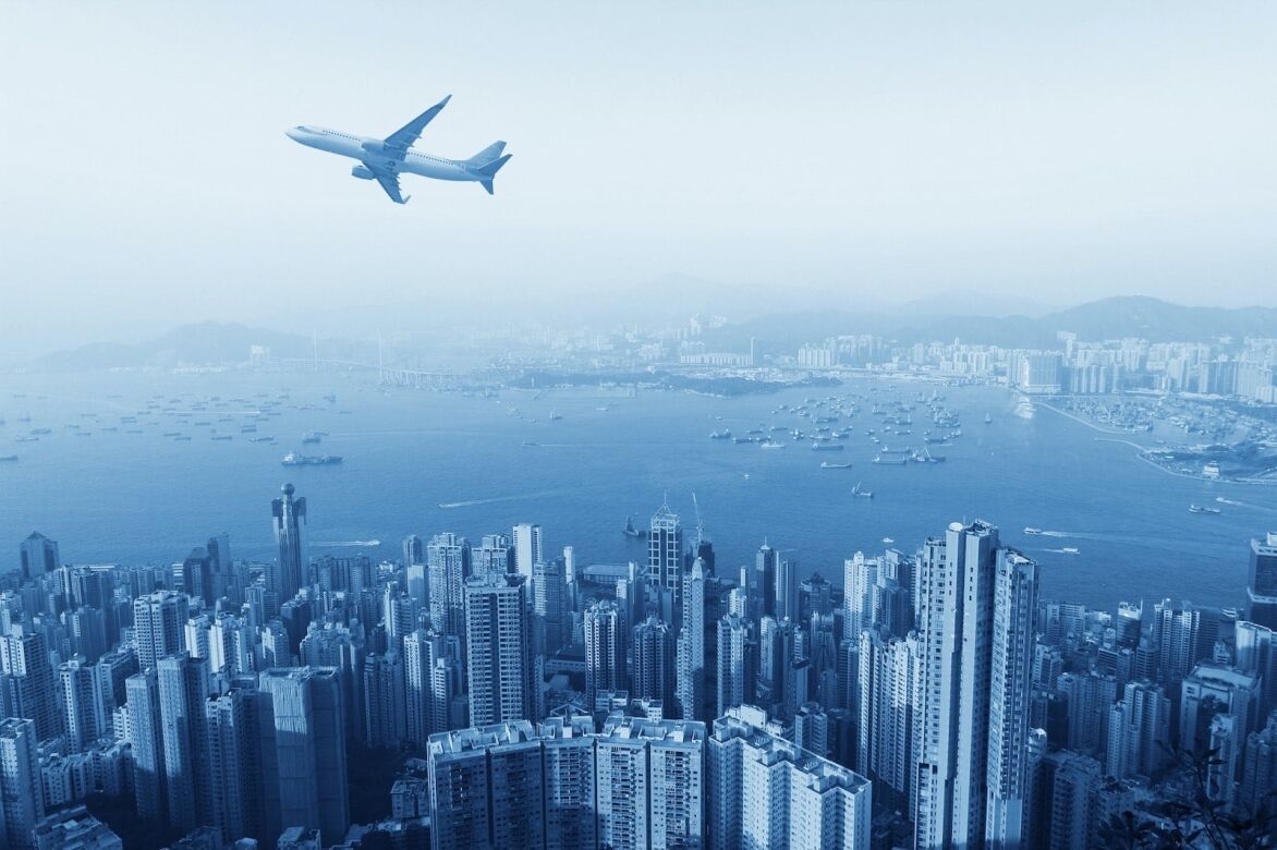 A panoramic view of a bustling city skyline with tall buildings, an airplane flying above, and a harbor filled with boats under a hazy blue sky.