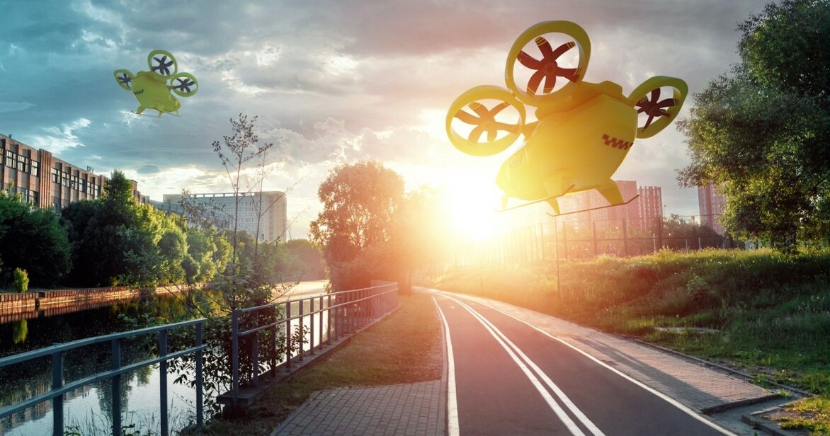 Two yellow drones fly above a tree-lined path beside a river during sunset, casting shadows against a vibrant sky.