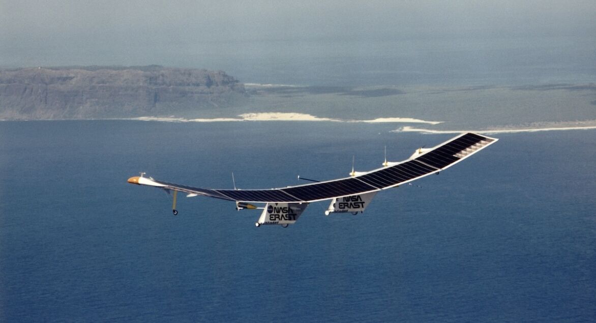 A large solar-powered aircraft glides above the ocean, with a mountainous coastline visible in the background under clear blue skies.