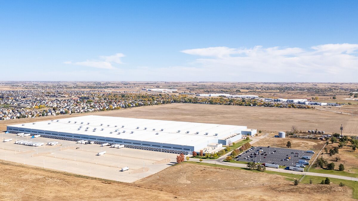 Aerial view of a large industrial warehouse surrounded by open land, with nearby residential areas and multiple parked vehicles.