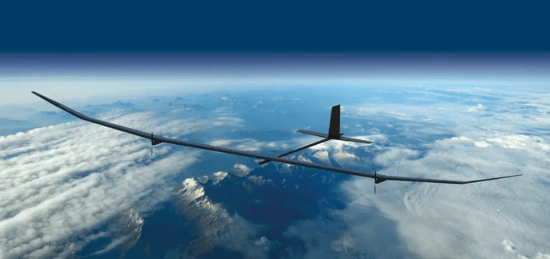 A sleek, elongated drone flies high above a cloud-covered landscape, showcasing its wingspan against a backdrop of blue sky and distant mountains.