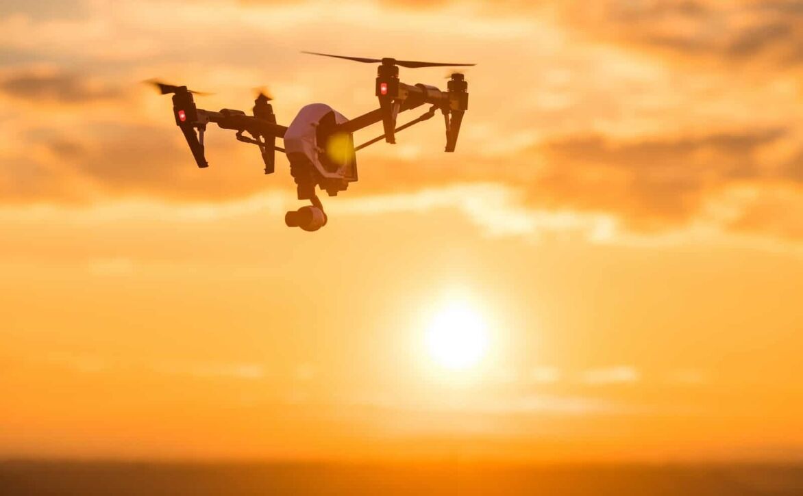 A drone flies against a vibrant sunset, silhouetted by warm hues of orange and yellow.