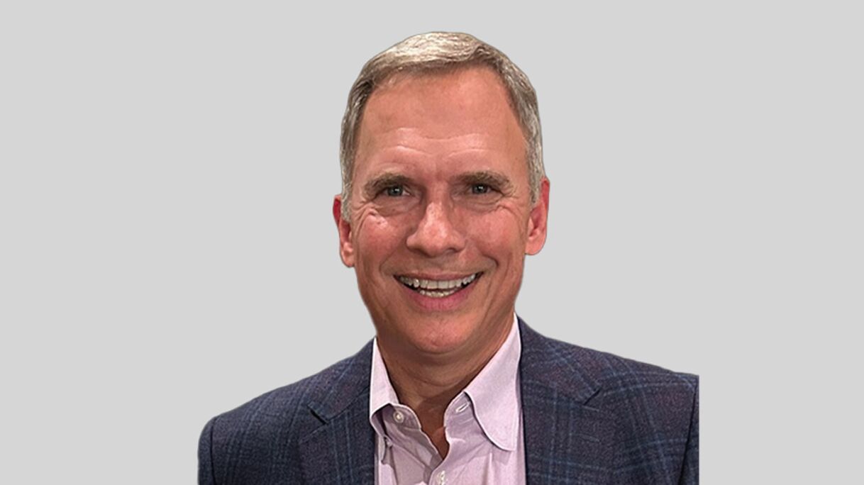 A smiling man with short hair, wearing a plaid blazer over a light shirt, poses against a grey background.