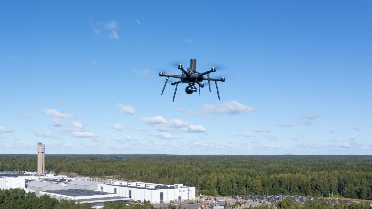 A drone hovers over a landscape with dense green forest and industrial buildings under a clear blue sky.