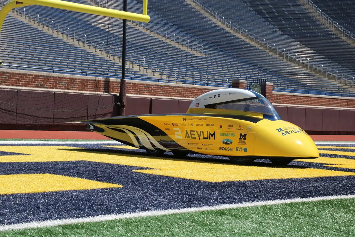 A sleek yellow vehicle with a streamlined design stands on a football field, surrounded by a stadium's empty seating.