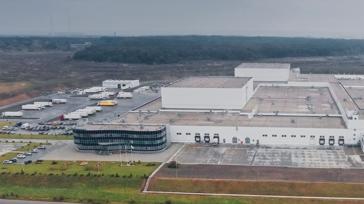 Aerial view of a large industrial facility with multiple loading docks and trucks in the parking area, surrounded by a forested landscape.