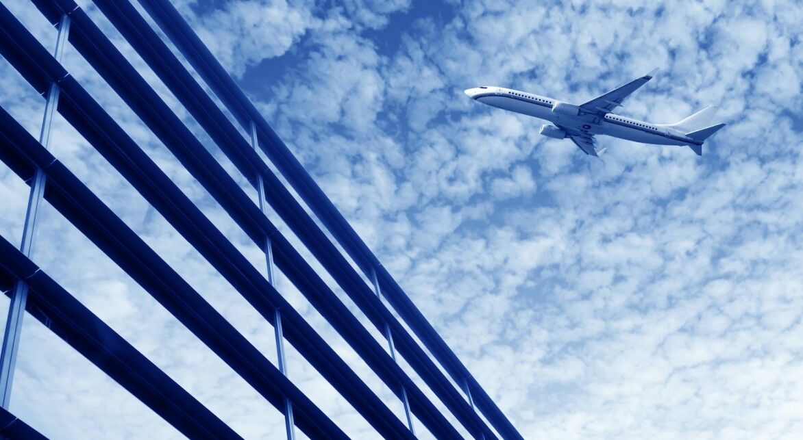 A modern building with reflective windows contrasts with a flying airplane against a blue sky filled with clouds.
